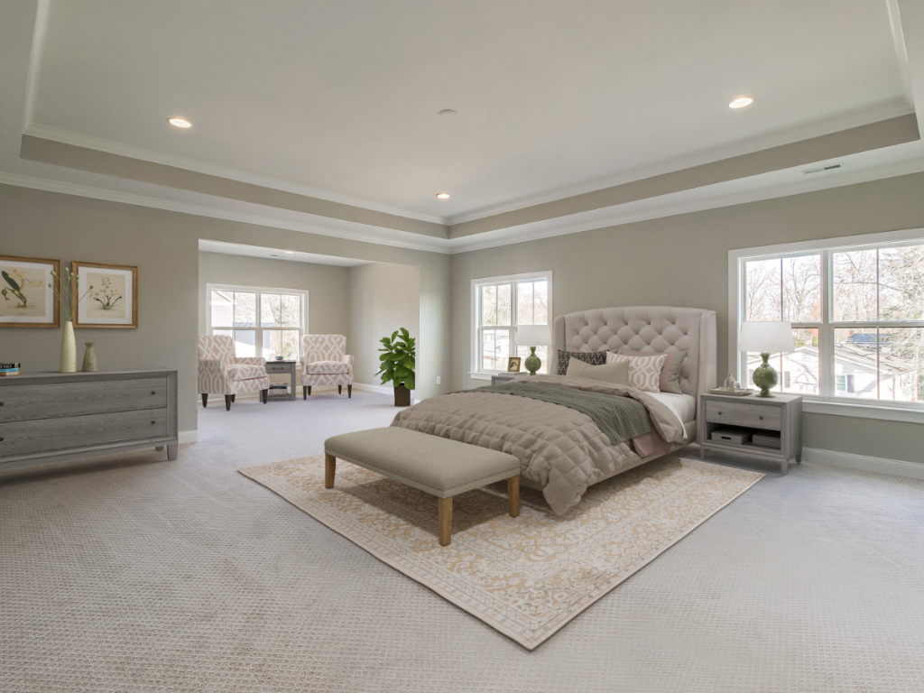 Owners suite bedroom. Soft white paint with recessed lighting. Bed in the middle of the room with a dresser on the left side. Sitting room in the back with a couch and a coffee table. 