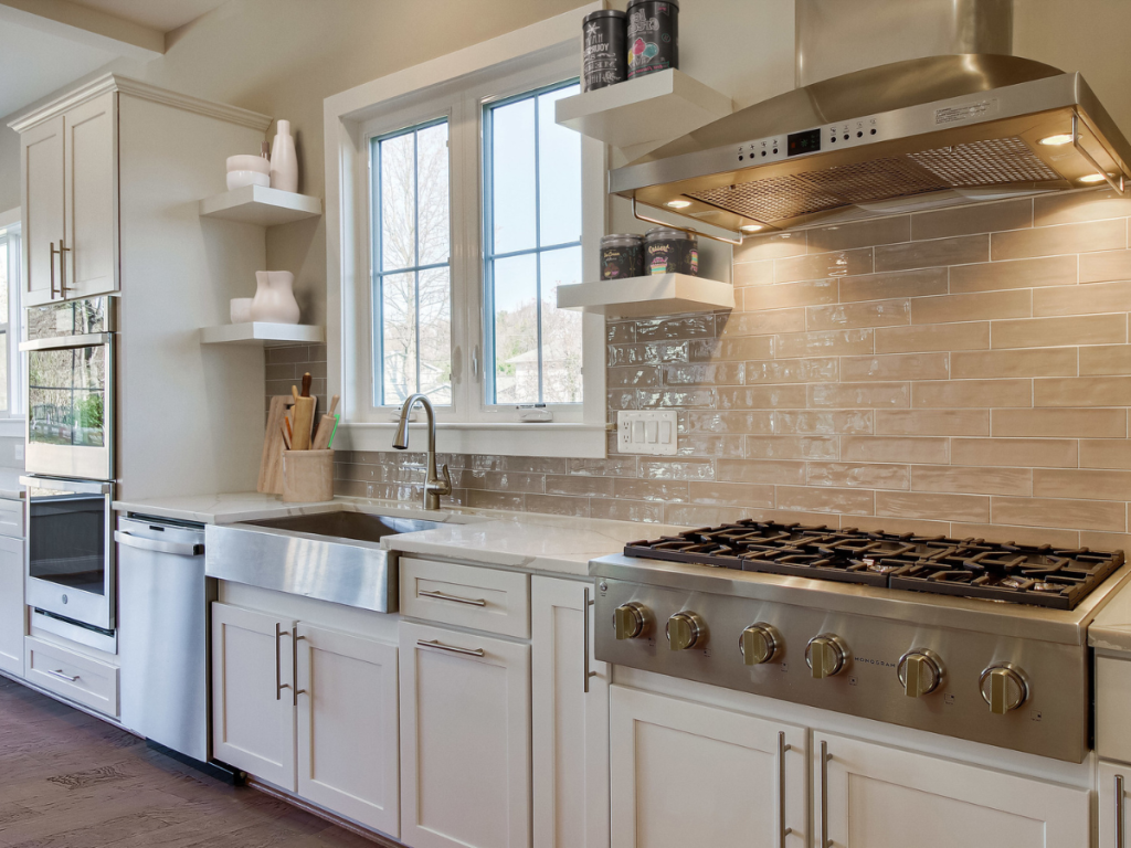 Close up of kitchen backsplash. Range and kitchen hood are stainless steel. Cabinetry below is white. Cabinetry near sink is floating shelves. 